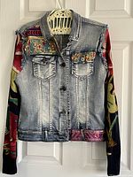 Front view of the Desigual women's blue jean jacket showing floral patchwork and knit textile sleeves with button closure.