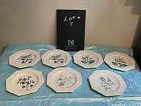 Seven octagonal luncheon plates arranged in two rows showcasing different herb illustrations on each plate with a neutral background