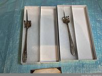 Two vintage novelty forks side by side in open white boxes on a blue textured surface, showing their distinctive curled prongs and decorative handles.