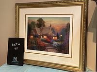 Framed serilithograph print titled "Evening Glow" showing a cozy cottage at sunset surrounded by colorful flowers and lit windows.