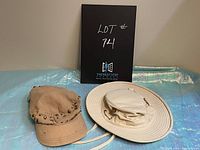 Two hats side by side on a light blue iridescent surface: a beige Le Chateau cap with rivets and a white Tilley Endurable sun hat with straps.