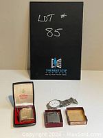 Two Ronson vintage lighters in their original cases and one vintage men's wristwatch all displayed on a white surface with a black lot sign behind.