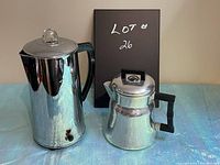 Two vintage metal coffee percolators on a blue surface with a black card labeled Lot #26 behind them; electric model is taller with clear lid knob and missing power cord, stove top model is smaller with square lid knob and angled handle.