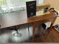 Photo showing the entire wooden sewing table with the sewing machine mounted and extension flap folded out, the metal foot pedal and the underside sewing machine metal bed visible.
