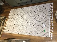 Full view of rectangular beige and off-white area rug with diamond pattern and fringed edges on hardwood floor.