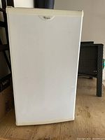 Closed front view of white Whirlpool bar fridge on wooden floor with wicker furniture in background