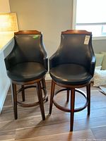 Two matching swivel stools made in Malaysia with black faux leather upholstery and wood frames, shown side by side.