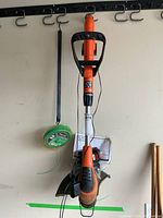 Full view of the Black & Decker wireless trimmer hanging against a wall with the loop handle, orange and black body, and green trimmer line visible.