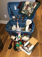 Blue tote filled with assorted Christmas decorations including plush dolls outside tote and quilted wall hanging over lid