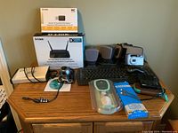 Lot contents on wooden table showing D-Link router and adapter boxes, speakers, Logitech keyboard and mouse, cameras, charger, and cables.