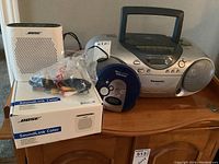 Photo showing Bose SoundLink Color Bluetooth speaker in white with its original box and cables, Panasonic RX-DT25 portable radio and CD player, and Koss DB85 Discman all placed on wooden furniture.