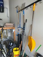 Garage wall with multiple garden tools including shovels, hoe, hand tiller, garden fork, and yellow bucket