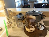 Photo showing full set of appliances on kitchen counter including crock pot, waffle maker, hand mixer on stand with beaters, food processor with blades, emulsifier, and electric kettle.