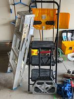 Four step aluminum ladder, three step stool, and folding moving cart grouped together in a garage environment.