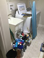 Photo of laundry room with washing machine, utility sink, two ironing boards (blue and white), and various bottles of cleaning liquids, rags and other supplies on floor.