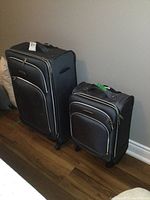 Two black luggage pieces with grey trim standing on wooden floor against a wall, showing full height and overall exterior design.
