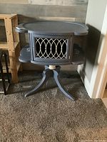 Full view of painted wood occasional table with central door and raised scalloped top edge, sitting on carpeted floor.