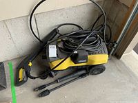 Full view of the yellow and black Karcher pressure sprayer with power cord and two spray wands lying in a garage.