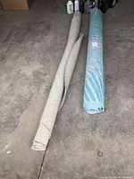 Two rolled rugs side by side on floor. One beige/grey outdoor mat and one blue patterned indoor/outdoor rug.