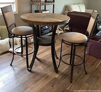 Full view of the round wooden bistro table with decorative top and metal base, alongside two matching swivel chairs with beige padded seats and backs in a living room setting.