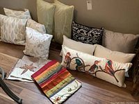 Photo of various throw cushions arranged on floor against wall, showing variety of designs and colors including floral, leaf, and bird embroidery.