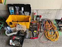 Plastic toolbox containing various hand tools, a B and D electric drill with cable, multiple extension cords coiled on the floor, and small containers with screws and cable ties.