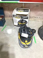 Front view of the Stinger wet/dry vacuum next to its original box showing the coiled hose on top and the yellow and grey body.