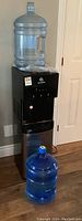 Full view of Avalon water cooler with clear jug on top and a blue jug in front on floor.
