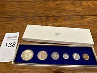 Photo showing six Canadian coins from 1967 Centennial year inside a white collection box with a blue velvet insert on wooden surface next to Maxsold lot tag.