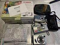 Photo of lot including Canon PowerShot A310 box with manuals, several compact digital cameras, and camera cases spread on a table.