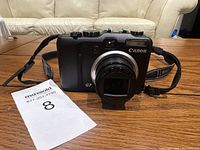 Front view of Canon PowerShot G7 camera on wood table with attached strap and lot number card.