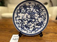 Front view of a round porcelain plate featuring a blue five-dragon design placed on a black display stand on a wooden surface.
