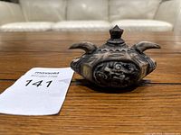 Mini decorative ink container displayed on wooden table with lot card number 141 visible. The container is small, ceramic or clay, with ornate carved floral patterns and a lid with a pointed knob.