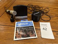 Photo of microphone on stand, wireless receiver, guitar string package, and a small black portable speaker with coiled cable on wooden surface.