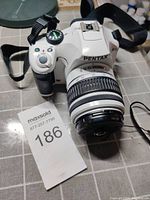 White Pentax DSLR camera angled view with lens attached and strap visible