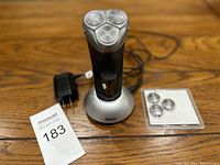 Philips electric rotary shaver standing on its charging base with attached power adapter and sealed replacement blades pack to the right, laid out on a wooden surface.