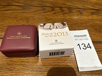 Maroon presentation box, outer packaging box with baby feet and text 'born in 2013', and auction lot tag 134 on wooden surface.