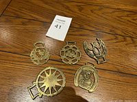 Photo of five vintage brass wall hangings on a wood floor, including horseshoe and other decorative motifs.