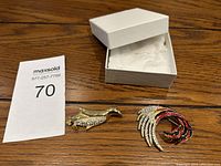 Top view of two vintage brooches; a gold-tone bird-shaped with rhinestones and a multi-colored wing design brooch beside an open white jewelry box.