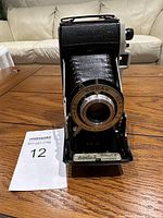 Front view of vintage Kodak Junior II camera showing the bellows and metal lens ring with shutter markings, resting on wooden surface.