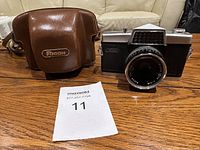 Front view of the Ricoh camera next to its brown leather Ricoh branded case, placed on a wooden surface.