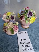 Three small pink vases with multiple porcelain flowers in purple, yellow, pink, and green colors.