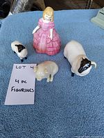 Four porcelain figurines on blue cloth, including a girl in a pink dress, a small pig, and two sheep figures.