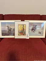Three framed prints pictured on a red couch: one Arbutus print in silver metal frame and two cowboy prints in gold frames, displayed side by side.