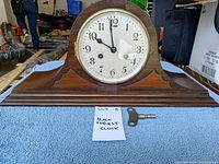 Wooden tabletop mantel clock with winding key and handwritten note "Blackforest Clock" on a blue cloth surface.