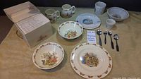 Collection of Bunnykins bone china pieces with Peter Rabbit characters along with four silverware spoons, displayed on cloth.