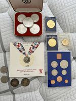 Assorted coins and medallions displayed on quilt including Olympic silver medallion set in case, Expo 86 medallions, Commonwealth Games £2 presentation, UK proof set, and loose coins.