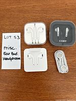 Four sets of earbud headphones placed on wooden surface, three Apple white earbuds in various packaging and one Coby earbud in black case.