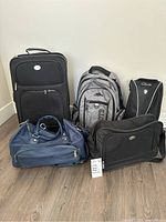 Photo of entire lot showing all bags: black wheeled suitcase, grey Roots backpack, blue duffel bag, black carry bag, and Clarks shoe bag.