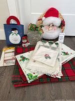 Photo of assorted Christmas linens, blue snowman gift bag, small red snowman ornament, clear glass cube decoration, and Santa Claus wreath.